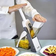 Person using a kitchen tool to slice vegetables on a table with food items around.