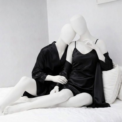 Two mannequins in black satin nightgowns sitting on a bed in a minimalistic room.