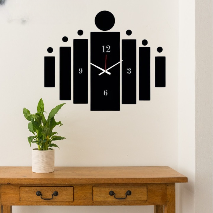 Black wall clock with unique design on a white wall above a wooden table with a plant.