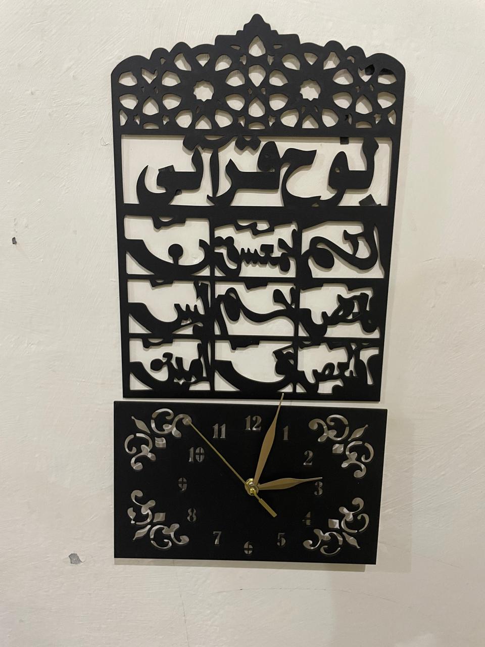 Black metal Islamic wall clock featuring Arabic calligraphy of Ayat al-Kursi and a square clock face with gold hands, mounted on a white wall.